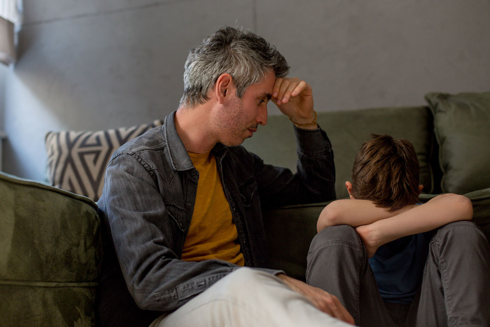 adolescent boy with head in arms depressed while father is confused on how to help